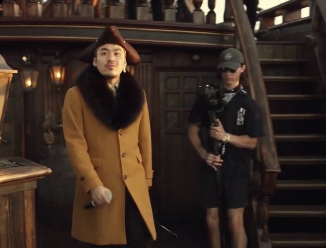 결과 영상 같은 순간. 삼각모와 털 깃 코트 차림의 인물이 나무로 된 선실 앞을 걷고, 같은 카메라맨이 같은 자리에 서 있다.